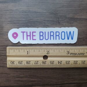 K&ouml;nnte beinhalten: Ein wei&szlig;er Aufkleber mit einem rosa Standort-Pin und dem Text "THE BURROW" in lila und blauen Buchstaben.