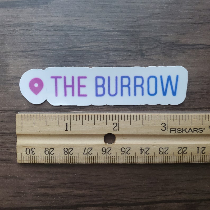 K&ouml;nnte beinhalten: Ein wei&szlig;er Aufkleber mit einem rosa Standort-Pin und dem Text "THE BURROW" in lila und blauen Buchstaben.