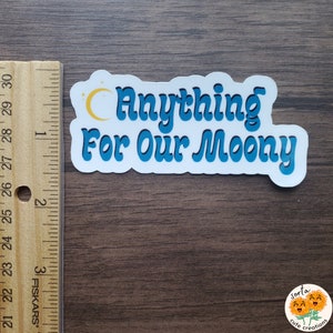 May include: A white sticker with the text "Anything For Our Moony" in blue, outlined in a lighter blue. A yellow crescent moon and stars are above the word "Anything". The sticker is on a wooden surface.