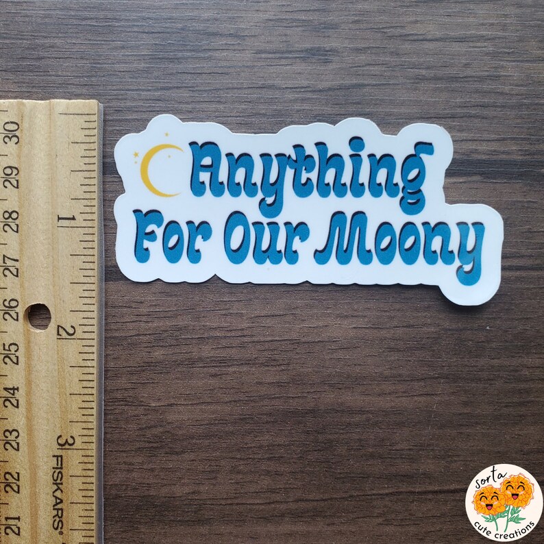 May include: A white sticker with the text "Anything For Our Moony" in blue, outlined in a lighter blue. A yellow crescent moon and stars are above the word "Anything". The sticker is on a wooden surface.