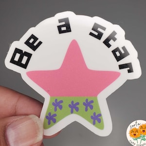 May include: A white sticker with the text "BE A STAR" in black block letters. The sticker features a pink star with a green band at the bottom adorned with purple flower shapes. The sticker has a unique, irregular shape.