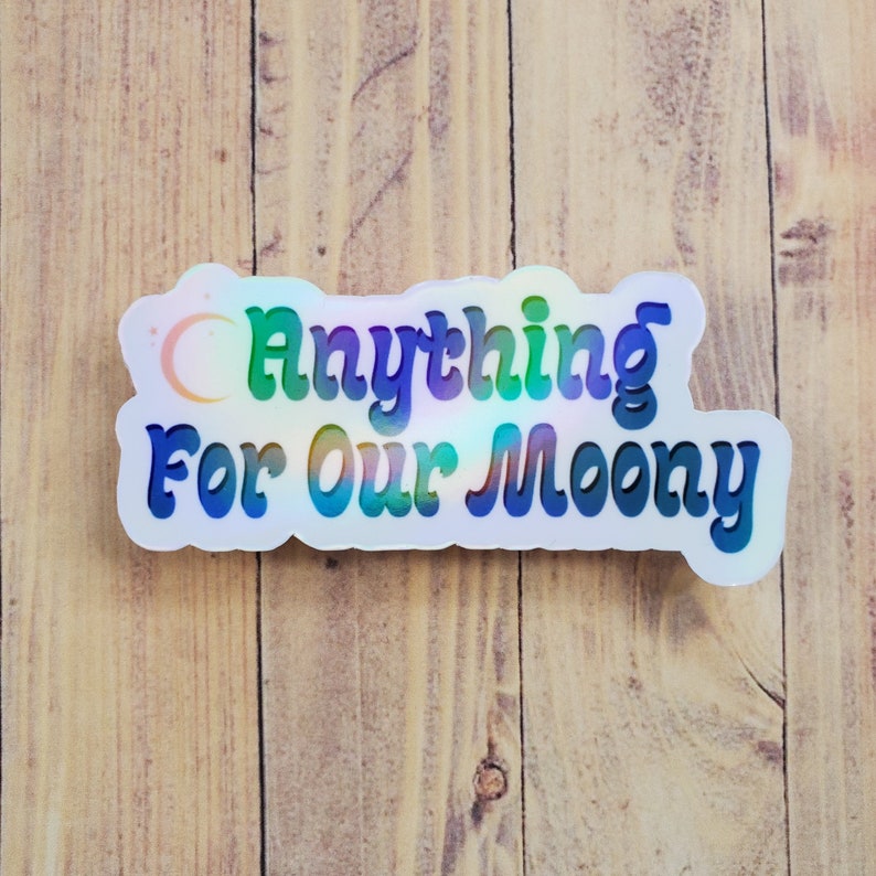 May include: A holographic sticker with the text "Anything For Our Moony" in a colorful, retro font. The sticker has a white border and a crescent moon design. The sticker is on a light brown wooden surface.
