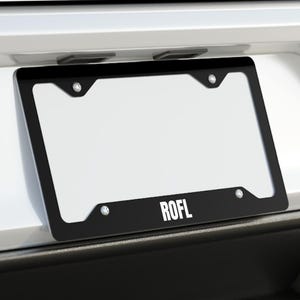 May include: Black license plate frame with the text "ROFL" on the bottom, mounted on the back of a white car. The frame has a white blank license plate. The car has dual exhaust pipes.