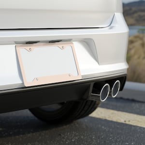 May include: A light pink license plate frame is mounted on the rear of a white car. The frame surrounds a blank license plate. Dual chrome exhaust pipes are visible below the bumper.