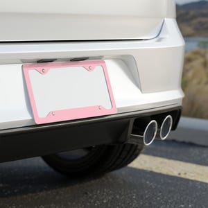 May include: A pink license plate frame is mounted on the back of a white car. The frame has a rectangular shape with rounded corners and is secured with four screws. The car's exhaust pipes are visible below the bumper.