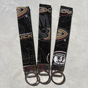 Może przedstawiać: Trzy czarne breloki do kluczy z logo Anaheim Ducks w kolorze złotym, białym i pomarańczowym. Każdy brelok ma srebrny brelok do kluczy. Tkanina przedstawia nazwę i logo drużyny.