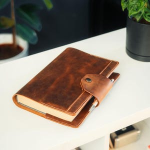 Könnte beinhalten: Ein braunes, ledergebundenes Tagebuch mit einem Stift, der in die Klappe gesteckt ist. Das Tagebuch liegt auf einem weißen Regal.