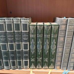 May include: A collection of fourteen hardcover books arranged on a shelf. Five dark green volumes are labeled D.H. Lawrence Complete Works, four green volumes are titled Masterpieces of Maupassant, and five grey volumes feature decorative gold-toned spines.