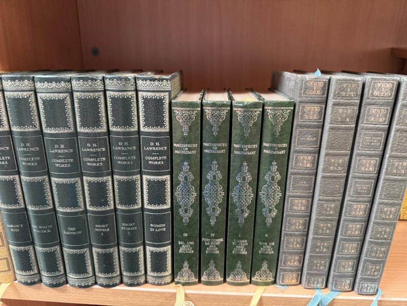 May include: A collection of fourteen hardcover books arranged on a shelf. Five dark green volumes are labeled D.H. Lawrence Complete Works, four green volumes are titled Masterpieces of Maupassant, and five grey volumes feature decorative gold-toned spines.