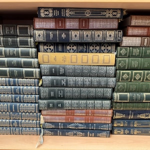 May include: A collection of hardcover books arranged on a wooden shelf. The books feature various colors including dark blue, green, red, and tan, with gold-toned decorative patterns and lettering on the spines. The books are stacked horizontally.