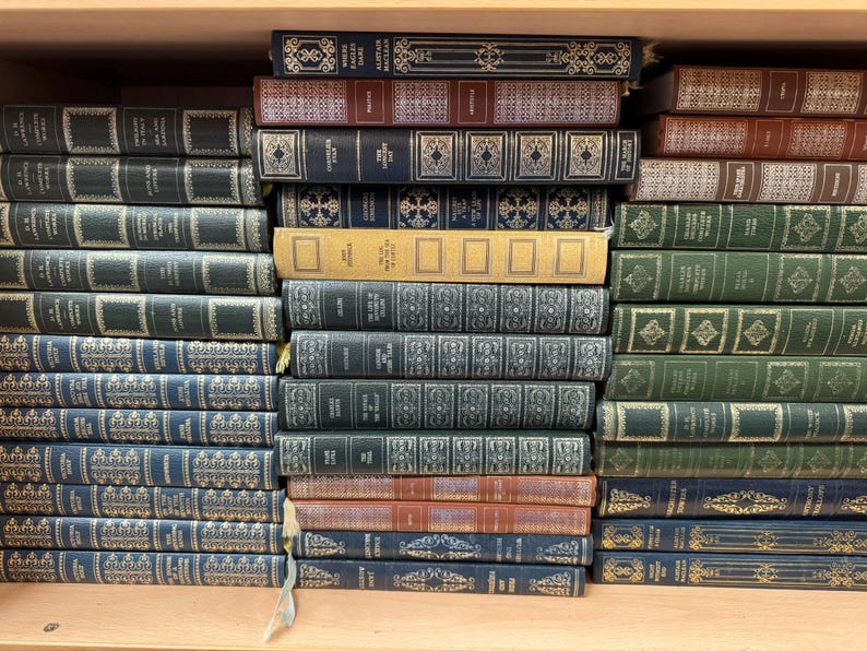 May include: A collection of hardcover books arranged on a wooden shelf. The books feature various colors including dark blue, green, red, and tan, with gold-toned decorative patterns and lettering on the spines. The books are stacked horizontally.