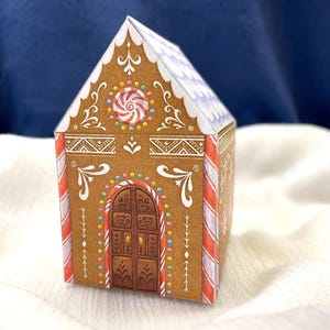 May include: A decorative gingerbread house box with brown walls, white icing details, and a candy cane striped roof. The front features a brown door and a red and white swirl candy. The box is on a white surface.