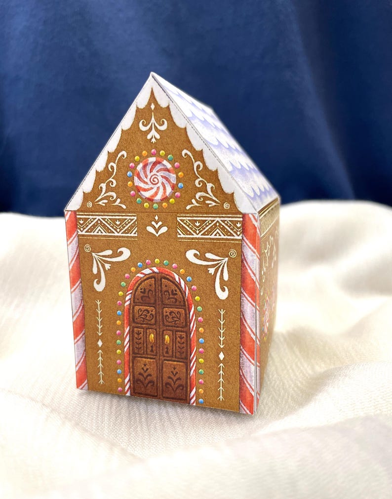 May include: A decorative gingerbread house box with brown walls, white icing details, and a candy cane striped roof. The front features a brown door and a red and white swirl candy. The box is on a white surface.