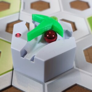 Puede incluir: Una pieza de juego hexagonal blanca con una palanca de pl&aacute;stico verde y una bola roja. La pieza est&aacute; en un tablero de juego hexagonal con azulejos verdes y blancos.