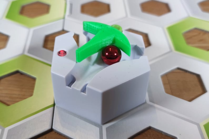 Puede incluir: Una pieza de juego hexagonal blanca con una palanca de pl&aacute;stico verde y una bola roja. La pieza est&aacute; en un tablero de juego hexagonal con azulejos verdes y blancos.