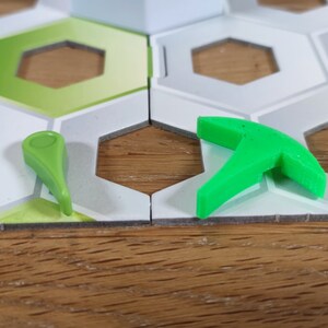 Puede incluir: Dos piezas de juego de pl&aacute;stico verde en un tablero de juego hexagonal blanco y gris. Una pieza es una peque&ntilde;a flecha puntiaguda. La otra pieza es una herramienta de tres puntas m&aacute;s grande.