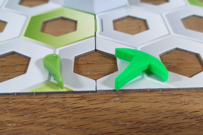 Puede incluir: Dos piezas de juego de pl&aacute;stico verde en un tablero de juego hexagonal blanco y gris. Una pieza es una peque&ntilde;a flecha puntiaguda. La otra pieza es una herramienta de tres puntas m&aacute;s grande.