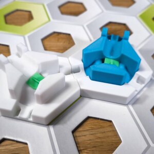 May include: Two game pieces on a white hexagonal game board. One piece is white with a green accent and the other is blue with a green accent.