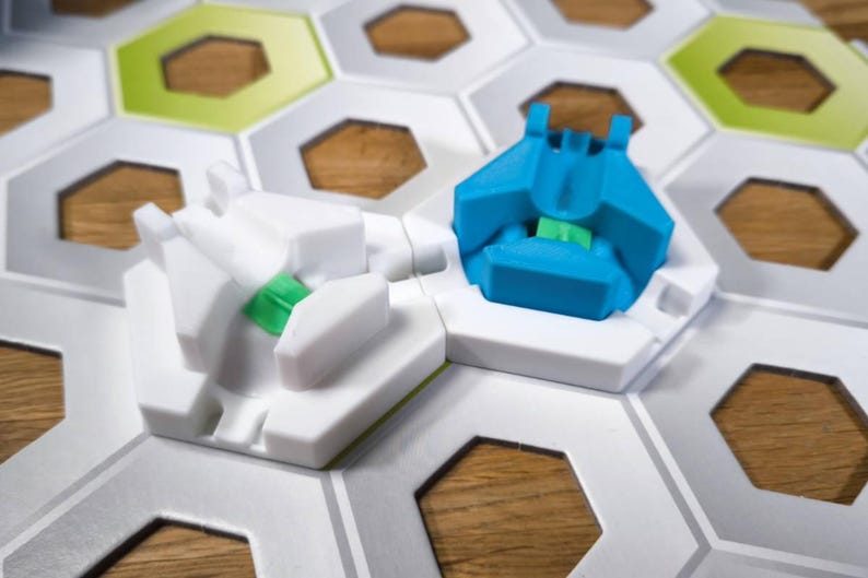 May include: Two game pieces on a white hexagonal game board. One piece is white with a green accent and the other is blue with a green accent.
