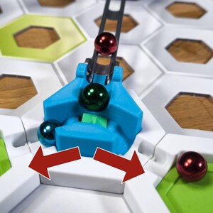 May include: A blue and white game board with a metal arm holding a green and red marble. The arm is moving the marble to the right. The board is made of hexagonal tiles.
