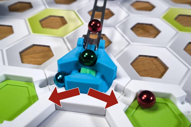 May include: A blue and white game board with a metal arm holding a green and red marble. The arm is moving the marble to the right. The board is made of hexagonal tiles.