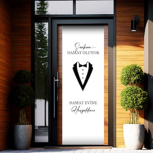 May include: A white door with a black tuxedo graphic and the text "Serkan DAMAT OLUYOR DAMAT EVINE Hosgeldiniz" in black. The door is framed by a black door frame and is set against a wooden wall.