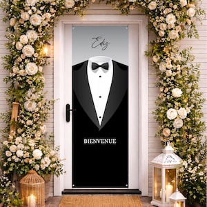 May include: A black and white door banner featuring a tuxedo design with a bow tie and the word "BIENVENUE" at the bottom. The banner has the name "Ediz" at the top. The door is decorated with white flowers.