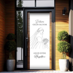 May include: A white door with a black frame and a white sign that says "Dilara Gelin Oluyor" and "Gelin Evinize Hosgeldiniz". The sign has a line drawing of a bride in a wedding dress.