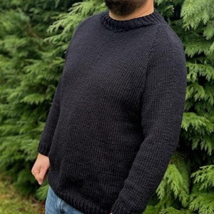 Puede incluir: Un suéter de punto azul marino oscuro con cuello redondo y mangas largas. El suéter tiene un patrón de punto texturizado y puños y dobladillo acanalados. La persona lleva vaqueros azules. El fondo es un arbusto verde y frondoso.