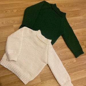 Könnte beinhalten: Zwei Strickpullover, einer in Creme und einer in Dunkelgrün. Beide Pullover sind gefaltet und auf einer Holzoberfläche ausgelegt.