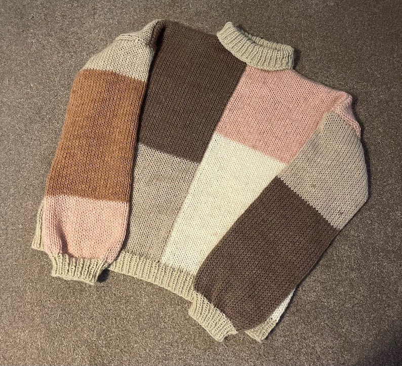 May include: A brown, pink, and beige knitted sweater with a patchwork design. The sweater has a high neck and long sleeves.