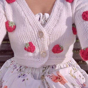 以下が含まれることがあります： Vネックとボタンが付いた白いニットカーディガン。カーディガンは刺繍されたイチゴで飾られています。
