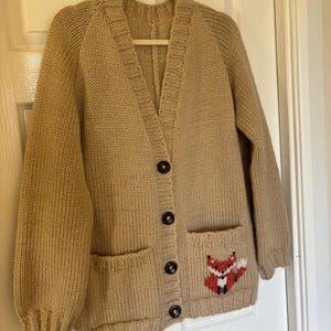 Może przedstawiać: Beżowy kardigan z dzianiny z haftowanym lisem na kieszeni. Kardigan ma dekolt w kształcie litery V i zapięcie na guziki.