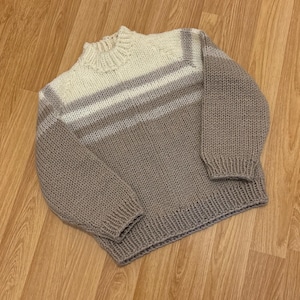 Puede incluir: Un suéter tejido a mano en tonos grises y crema. El suéter presenta un cuello, puños y dobladillo acanalados. Rayas horizontales en diferentes tonos de gris adornan la parte superior del pecho. Las mangas son largas y el estilo es informal.