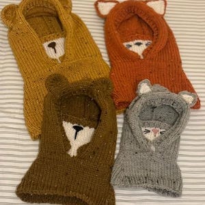 May include: Four knitted animal hoods in gold, orange, brown, and grey. Each hood features a different animal face: bear, fox, bear, and cat. The hoods have ear details and embroidered facial features.