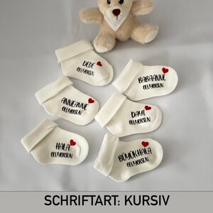 K&ouml;nnte beinhalten: Cremefarbene Babysocken mit schwarzem Text und einem roten Herz. Jede Socke hat einen anderen Namen, darunter "DEDE", "BABAANNE", "ANNEANNE", "DAYI", "HALA" und "B&Uuml;Y&Uuml;KHALA". Ein kleiner Teddyb&auml;r ist im Hintergrund. Der Text "SCHRIFTART: KURSIV" steht unten.