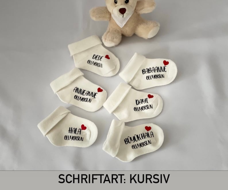 K&ouml;nnte beinhalten: Cremefarbene Babysocken mit schwarzem Text und einem roten Herz. Jede Socke hat einen anderen Namen, darunter "DEDE", "BABAANNE", "ANNEANNE", "DAYI", "HALA" und "B&Uuml;Y&Uuml;KHALA". Ein kleiner Teddyb&auml;r ist im Hintergrund. Der Text "SCHRIFTART: KURSIV" steht unten.