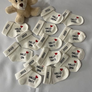 K&ouml;nnte beinhalten: Eine Ansammlung wei&szlig;er Babysocken, jeweils mit dem Jahr "2023" auf dem B&uuml;ndchen gestickt. Jede Socke hat ein rotes Herz und einen deutschen Gru&szlig; wie "Hallo Opa" oder "Hallo Oma". Ein kleiner Teddyb&auml;r ist im Hintergrund.