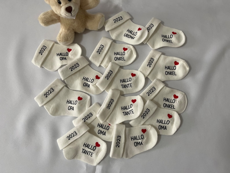K&ouml;nnte beinhalten: Eine Ansammlung wei&szlig;er Babysocken, jeweils mit dem Jahr "2023" auf dem B&uuml;ndchen gestickt. Jede Socke hat ein rotes Herz und einen deutschen Gru&szlig; wie "Hallo Opa" oder "Hallo Oma". Ein kleiner Teddyb&auml;r ist im Hintergrund.