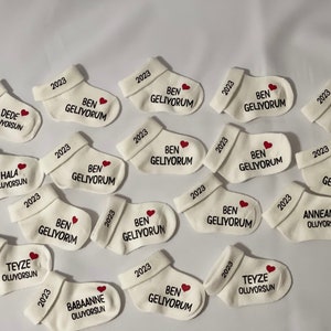 K&ouml;nnte beinhalten: Wei&szlig;e Babysocken mit gesticktem Text und einem roten Herz. Jede Socke hat "2023" auf dem B&uuml;ndchen gestickt. Einige Socken sagen "BEN GELIYORUM" mit einem Herz, andere haben S&auml;tze wie "DEDE OLUYORSUN".