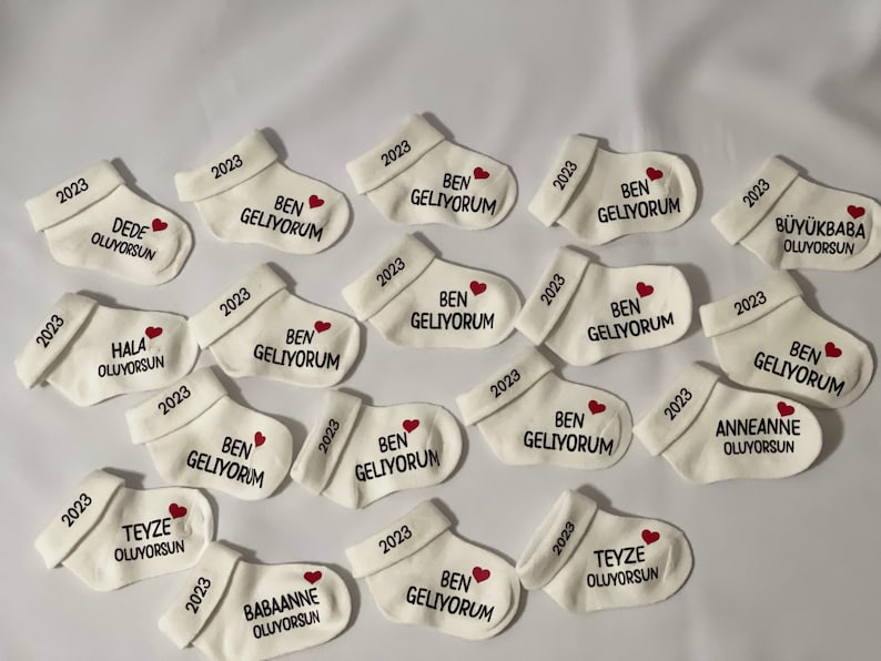 K&ouml;nnte beinhalten: Wei&szlig;e Babysocken mit gesticktem Text und einem roten Herz. Jede Socke hat "2023" auf dem B&uuml;ndchen gestickt. Einige Socken sagen "BEN GELIYORUM" mit einem Herz, andere haben S&auml;tze wie "DEDE OLUYORSUN".