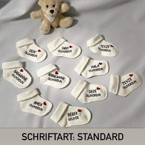 K&ouml;nnte beinhalten: Eine Ansammlung wei&szlig;er Babysocken mit t&uuml;rkischem Text und einem roten Herz. Die Socken sind um einen kleinen Teddyb&auml;ren auf einer wei&szlig;en Oberfl&auml;che angeordnet. Der Text "SCHRIFTART: STANDARD" befindet sich unten.