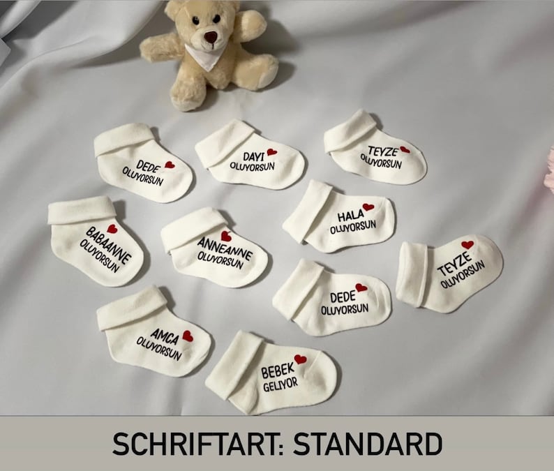 K&ouml;nnte beinhalten: Eine Ansammlung wei&szlig;er Babysocken mit t&uuml;rkischem Text und einem roten Herz. Die Socken sind um einen kleinen Teddyb&auml;ren auf einer wei&szlig;en Oberfl&auml;che angeordnet. Der Text "SCHRIFTART: STANDARD" befindet sich unten.