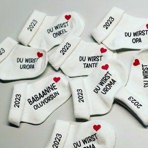 K&ouml;nnte beinhalten: Wei&szlig;e Babysocken mit schwarzem Text und einem roten Herz. Jede Socke hat das Jahr "2023" auf dem B&uuml;ndchen aufgedruckt. Die Socken sind mit S&auml;tzen wie "DU WIRST ONKEL" und "DU WIRST OPA" bedruckt.