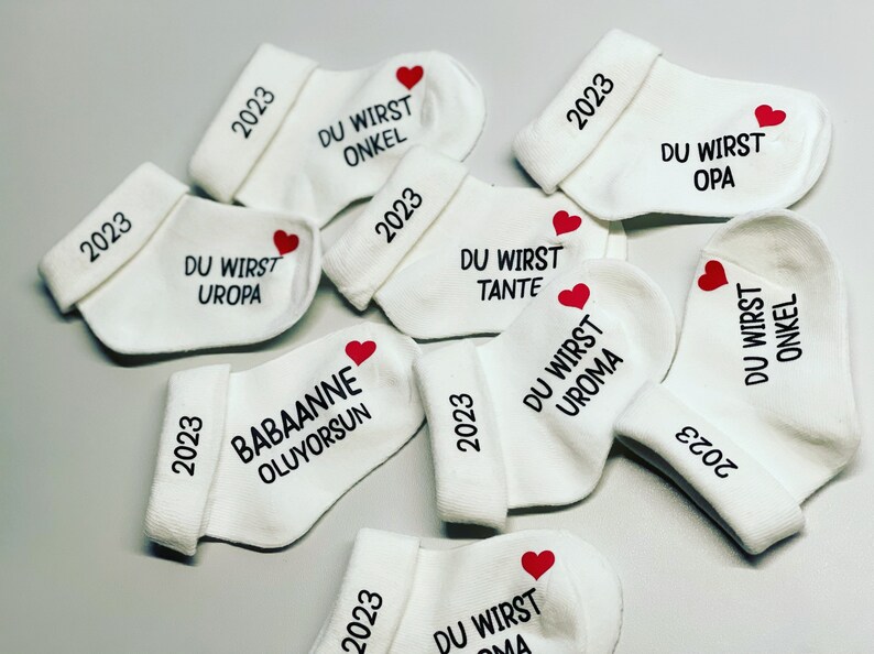 K&ouml;nnte beinhalten: Wei&szlig;e Babysocken mit schwarzem Text und einem roten Herz. Jede Socke hat das Jahr "2023" auf dem B&uuml;ndchen aufgedruckt. Die Socken sind mit S&auml;tzen wie "DU WIRST ONKEL" und "DU WIRST OPA" bedruckt.