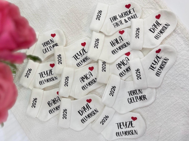 Könnte beinhalten: Weiße Babysocken mit schwarzem Text und roten Herzakzenten. Jede Socke trägt das Jahr "2026" sowie verschiedene türkische Phrasen. Ein charmantes und personalisiertes Geschenk für eine Babyparty