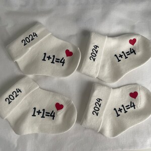 K&ouml;nnte beinhalten: Vier wei&szlig;e Babysocken mit Umschl&auml;gen, jeweils mit dem Jahr "2024", der Gleichung "1+1=4" und einem roten Herz bedruckt. Die Socken liegen auf einer wei&szlig;en Oberfl&auml;che.