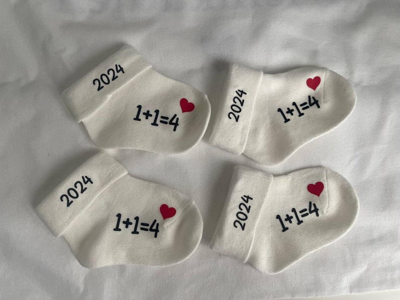 K&ouml;nnte beinhalten: Vier wei&szlig;e Babysocken mit Umschl&auml;gen, jeweils mit dem Jahr "2024", der Gleichung "1+1=4" und einem roten Herz bedruckt. Die Socken liegen auf einer wei&szlig;en Oberfl&auml;che.