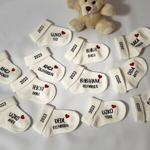K&ouml;nnte beinhalten: Eine Sammlung wei&szlig;er Babysocken, jede mit Namen und dem Jahr 2023 in schwarzer Schrift personalisiert. Ein kleines rotes Herz ist neben einigen Namen aufgedruckt. Ein Teddyb&auml;r ist im Hintergrund zu sehen.