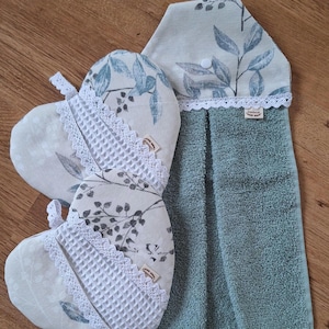 Könnte beinhalten: Ein Küchenset mit zwei herzförmigen Ofenhandschuhen und einem Hängehandtuch. Die Handschuhe und der Handtuchhalter haben einen hellbeigen Hintergrund mit einem blau-grauen Blättermuster, akzentuiert mit weißer Spitzenborte.